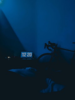 A dimly lit room with a blue color tone features a digital clock displaying 12:20. A bicycle is leaning against the wall, partially visible. The overall lighting creates a calm, subdued atmosphere.