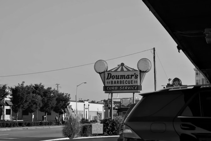 A black and white photograph of a vintage-looking signage for Doumar's Barbecue with curb service, situated in an urban setting. The surrounding area includes a street with cars and some trees. A fast-food sign is visible in the background.