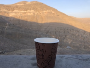 A paper cup with a decorative pattern is placed on a stone surface with a vast and rugged mountain landscape in the background. The mountain ranges show different shades of brown under the clear blue sky, indicating a dry and arid environment.
