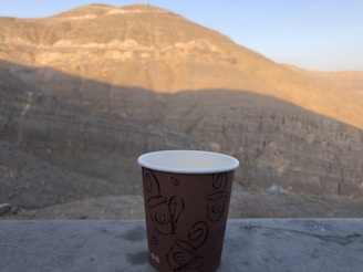 A paper cup with a decorative pattern is placed on a stone surface with a vast and rugged mountain landscape in the background. The mountain ranges show different shades of brown under the clear blue sky, indicating a dry and arid environment.
