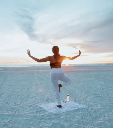 A person practicing a yoga pose on the beach during sunset. They stand on a mat with one leg raised and arms outstretched. The sun sets over the ocean, casting a warm glow and creating a serene atmosphere.