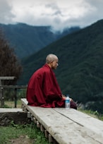 man in red robe sitting on brown wooden bench looking at green mountains during daytime