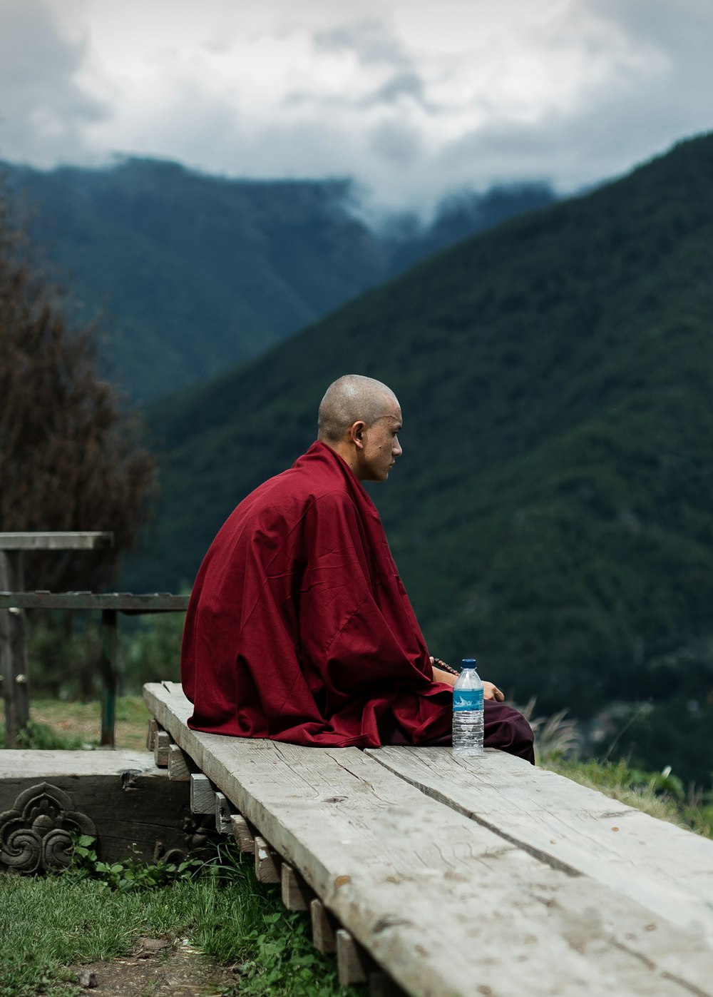 man in red robe sitting on brown wooden bench looking at green mountains during daytime