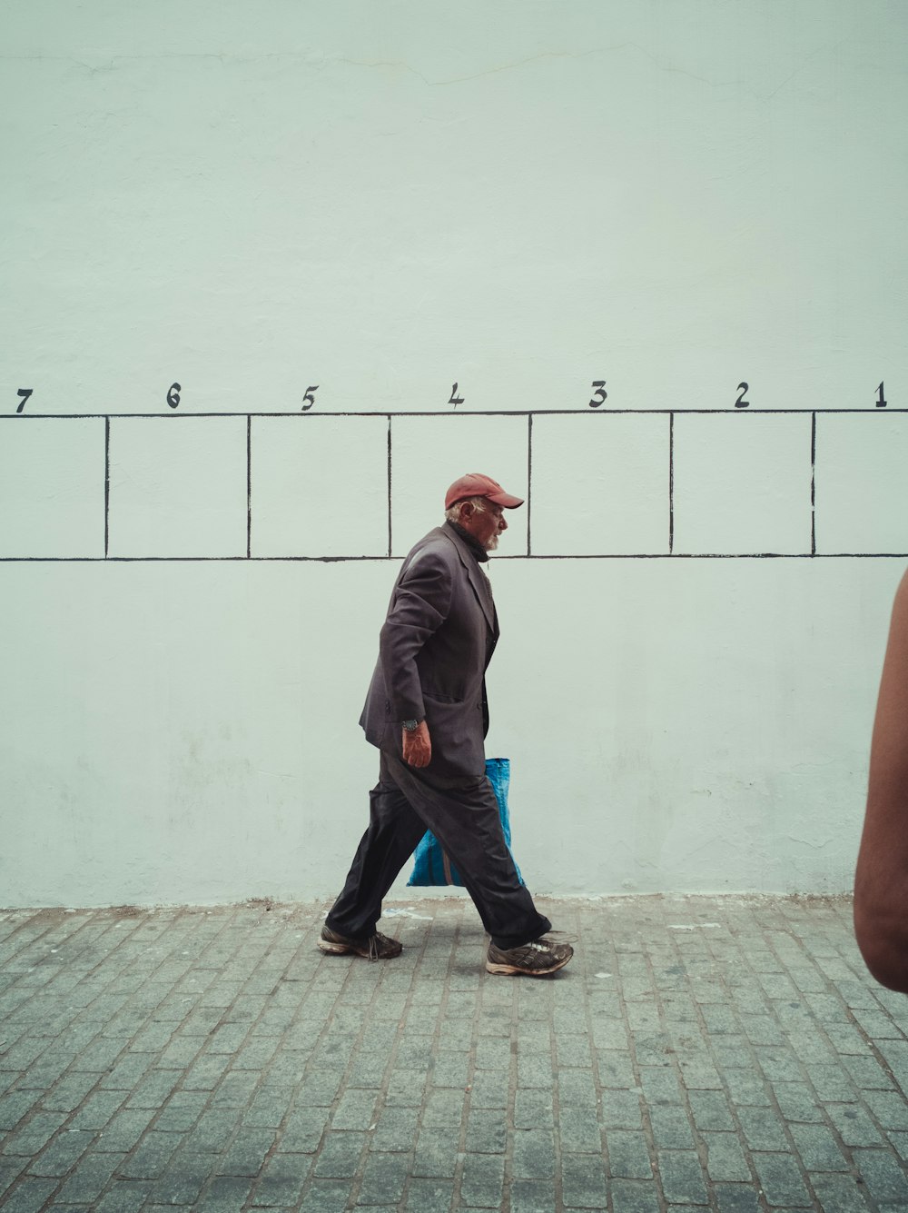 man in gray jacket and blue denim jeans standing beside white wall during daytime