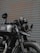 A black motorcycle is parked in front of a corrugated metal door. The bike features a round headlight, a rearview mirror with a logo, and a visible license plate. Details of the suspension and exhaust system are noticeable, and the paint on the vehicle appears to be well-maintained.