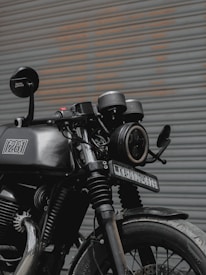 A black motorcycle is parked in front of a corrugated metal door. The bike features a round headlight, a rearview mirror with a logo, and a visible license plate. Details of the suspension and exhaust system are noticeable, and the paint on the vehicle appears to be well-maintained.