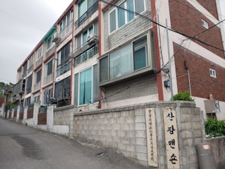 A multi-story residential building with a series of windows and balconies is situated on a slight slope. The building features a mix of materials, including brick and cement, with visible air conditioning units attached. A street runs alongside the building, and a wall with signs in a foreign script is present in the foreground.