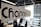 A modern office setting with a focus on a glass partition displaying the word 'Channel' printed in large black letters and 'lead' in lighter green letters. The background shows a blurred view of desks, papers, computers, office chairs, and vertical blinds, indicating an active work environment.