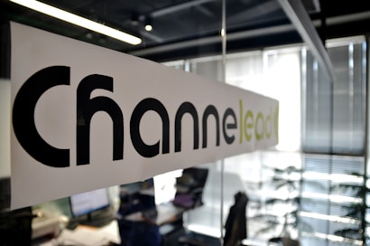 A modern office setting with a focus on a glass partition displaying the word 'Channel' printed in large black letters and 'lead' in lighter green letters. The background shows a blurred view of desks, papers, computers, office chairs, and vertical blinds, indicating an active work environment.