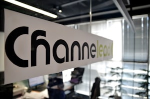 A modern office setting with a focus on a glass partition displaying the word 'Channel' printed in large black letters and 'lead' in lighter green letters. The background shows a blurred view of desks, papers, computers, office chairs, and vertical blinds, indicating an active work environment.