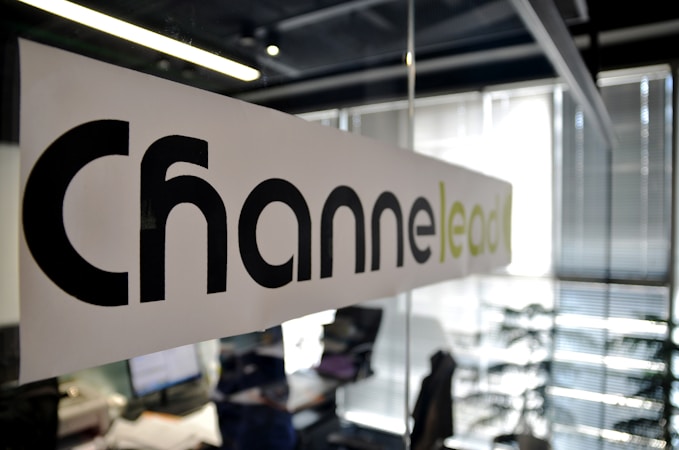 A modern office setting with a focus on a glass partition displaying the word 'Channel' printed in large black letters and 'lead' in lighter green letters. The background shows a blurred view of desks, papers, computers, office chairs, and vertical blinds, indicating an active work environment.