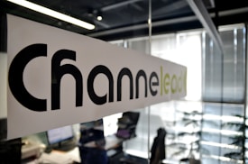 A modern office setting with a focus on a glass partition displaying the word 'Channel' printed in large black letters and 'lead' in lighter green letters. The background shows a blurred view of desks, papers, computers, office chairs, and vertical blinds, indicating an active work environment.