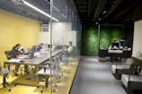 A modern office setting featuring a glass-walled conference room on the left with several individuals working at a long table, equipped with laptops and monitors. The room has a yellow theme with ergonomic chairs. Adjacent is a reception area with a dark, modern desk, where a person is working. The backdrop features a green, plant-covered wall, enhancing the aesthetics of the space.