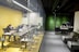A modern office setting featuring a glass-walled conference room on the left with several individuals working at a long table, equipped with laptops and monitors. The room has a yellow theme with ergonomic chairs. Adjacent is a reception area with a dark, modern desk, where a person is working. The backdrop features a green, plant-covered wall, enhancing the aesthetics of the space.