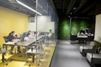 A modern office setting featuring a glass-walled conference room on the left with several individuals working at a long table, equipped with laptops and monitors. The room has a yellow theme with ergonomic chairs. Adjacent is a reception area with a dark, modern desk, where a person is working. The backdrop features a green, plant-covered wall, enhancing the aesthetics of the space.