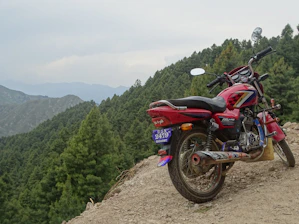 A rugged motorcycle parked on a dirt trail surrounded by dense forest and mountains.