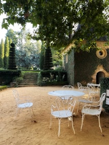 A serene garden setting with wrought iron chairs and a table on a sandy surface. The background features lush, green topiary trees and steps leading to a higher level. An ivy-covered stone building with a circular window adds a rustic touch, and the scene is shaded by overhanging tree branches.