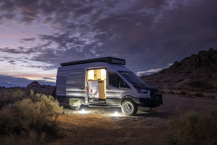 A campsite at dusk with a van, a folding chair, and a small camp stove set up outside