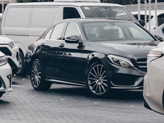 A parked black luxury sedan amidst several other cars, including a white SUV and a white van. The vehicles are aligned closely together in an urban setting, suggesting a busy street or parking lot scene.