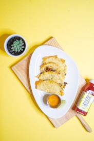 A wooden cutting board holds a white plate with three tacos, each filled with ingredients such as cheese and vegetables. A small cup of orange sauce and a lime wedge are on the plate. To the right, a bottle of red sauce with a yellow label is placed. In the top left corner, a small potted succulent adds a touch of greenery. The background is bright yellow, enhancing the vibrant presentation.