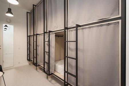 A room with multiple bunk beds, each enclosed with gray curtains for privacy. The beds are structured with black metal frames, and ladders are attached to access the upper bunks. The walls are white, and there's a full-length mirror on one wall. Ceiling lights with black shades provide illumination.