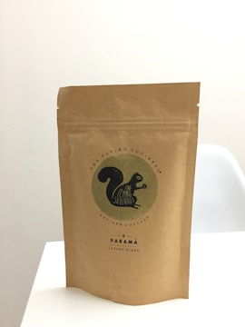 A brown paper bag stands upright on a white surface. The bag features a logo with an image of a black squirrel and the text 'The Flying Squirrel Artisan Coffee' along with 'Parama Luxury Blend'. The design is minimalistic and conveys a sense of artisan quality.