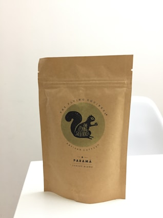 A brown paper bag stands upright on a white surface. The bag features a logo with an image of a black squirrel and the text 'The Flying Squirrel Artisan Coffee' along with 'Parama Luxury Blend'. The design is minimalistic and conveys a sense of artisan quality.