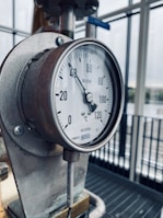 A close-up view of an industrial thermometer with a dial displaying temperatures from 0 to 120 degrees Celsius. The thermometer is mounted on a metallic structure, with a window and bars visible in the background.