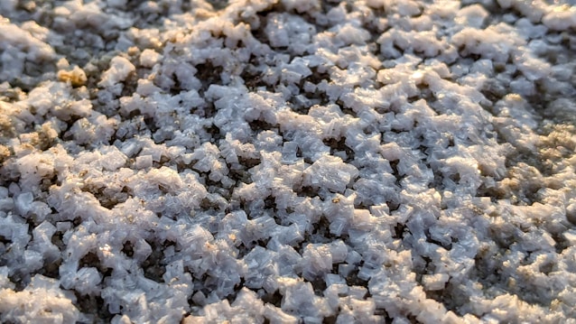Close-up view of salt crystals clustered densely, reflecting light in whites and soft grays. The texture is rough and granular, with the sunlight creating a slight shimmer across the uneven surface.