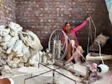 A woman is seated indoors, operating a traditional spinning wheel. She is surrounded by large bundles of yarn and raw fibers, which are piled on the floor. The room is enclosed by a rustic brick wall, and the woman wears colorful traditional clothing. The setting reflects a focus on labor and craftsmanship.