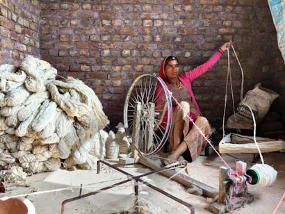 A woman is seated indoors, operating a traditional spinning wheel. She is surrounded by large bundles of yarn and raw fibers, which are piled on the floor. The room is enclosed by a rustic brick wall, and the woman wears colorful traditional clothing. The setting reflects a focus on labor and craftsmanship.