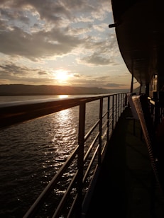 The image depicts a serene sunset view from the deck of a ship. The sun is low in the sky, casting a golden glow over the water. The ship's railing and part of the side structure are visible, with the horizon showing distant mountains and a sky filled with scattered clouds.