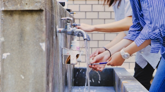 Several individuals are washing their hands under running water from outdoor faucets mounted on a concrete basin. The people are casually dressed, and one can be seen wearing a blue striped shirt. The setting appears to be outside, as indicated by the brick wall in the background.