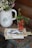A rustic setting featuring a white pitcher with floral design holding a bouquet of white daisies and purple wildflowers. Nearby, a glass filled with fresh strawberries is placed on top of a pile of illustration cards. A pair of scissors is visible on the cards, all arranged on a weathered wooden table. The scene exudes a natural and homely vibe.