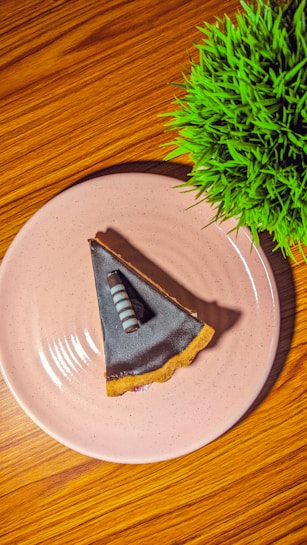 A slice of chocolate tart is served on a pale pink plate, positioned on a wooden surface. The tart features a layer of glossy dark chocolate, accentuated with a striped chocolate decoration on top. To the side, a vibrant green plant adds a touch of color to the composition.
