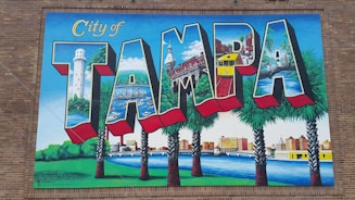 A vibrant mural featuring the word 'Tampa' with each letter depicting different landmarks and scenes from the city, such as a lighthouse, historic buildings, palm trees, a bridge, and a river. The mural is set against a brick wall and includes vivid colors and detailed illustrations.