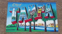 A vibrant mural featuring the word 'Tampa' with each letter depicting different landmarks and scenes from the city, such as a lighthouse, historic buildings, palm trees, a bridge, and a river. The mural is set against a brick wall and includes vivid colors and detailed illustrations.