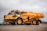 A large, heavy-duty yellow dump truck is positioned on a paved surface, carrying a load of material in its bed. The truck is branded with Volvo and appears to be used in construction or mining. The background features a cloudy sky and some sparse vegetation.