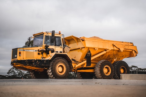A large, heavy-duty yellow dump truck is positioned on a paved surface, carrying a load of material in its bed. The truck is branded with Volvo and appears to be used in construction or mining. The background features a cloudy sky and some sparse vegetation.