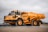 A large, heavy-duty yellow dump truck is positioned on a paved surface, carrying a load of material in its bed. The truck is branded with Volvo and appears to be used in construction or mining. The background features a cloudy sky and some sparse vegetation.