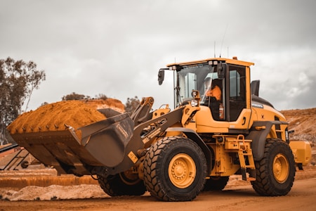 A yellow construction vehicle, specifically a wheel loader, is carrying a load of sand in its bucket. It is positioned on a dirt construction site with a cloudy sky overhead. Surrounding the vehicle are other construction elements, and a person is visible inside the cab operating the loader.