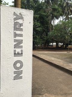 A white concrete post with the words 'NO ENTRY' written vertically in bold black letters is prominently shown. The post is situated next to a paved pathway in an outdoor environment, with lush green trees in the background.