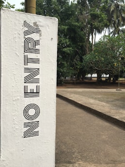 A white concrete post with the words 'NO ENTRY' written vertically in bold black letters is prominently shown. The post is situated next to a paved pathway in an outdoor environment, with lush green trees in the background.