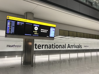 A modern airport terminal with a large sign for International Arrivals. There are digital display screens showing flight information, and directional signage pointing to Terminal 5 with directions for North and South exits. The environment is clean and spacious, with a tiled floor and overhead lighting.