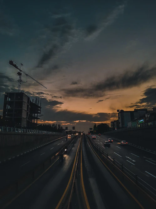 A wide highway under construction with heavy machinery and workers in safety gear during sunset