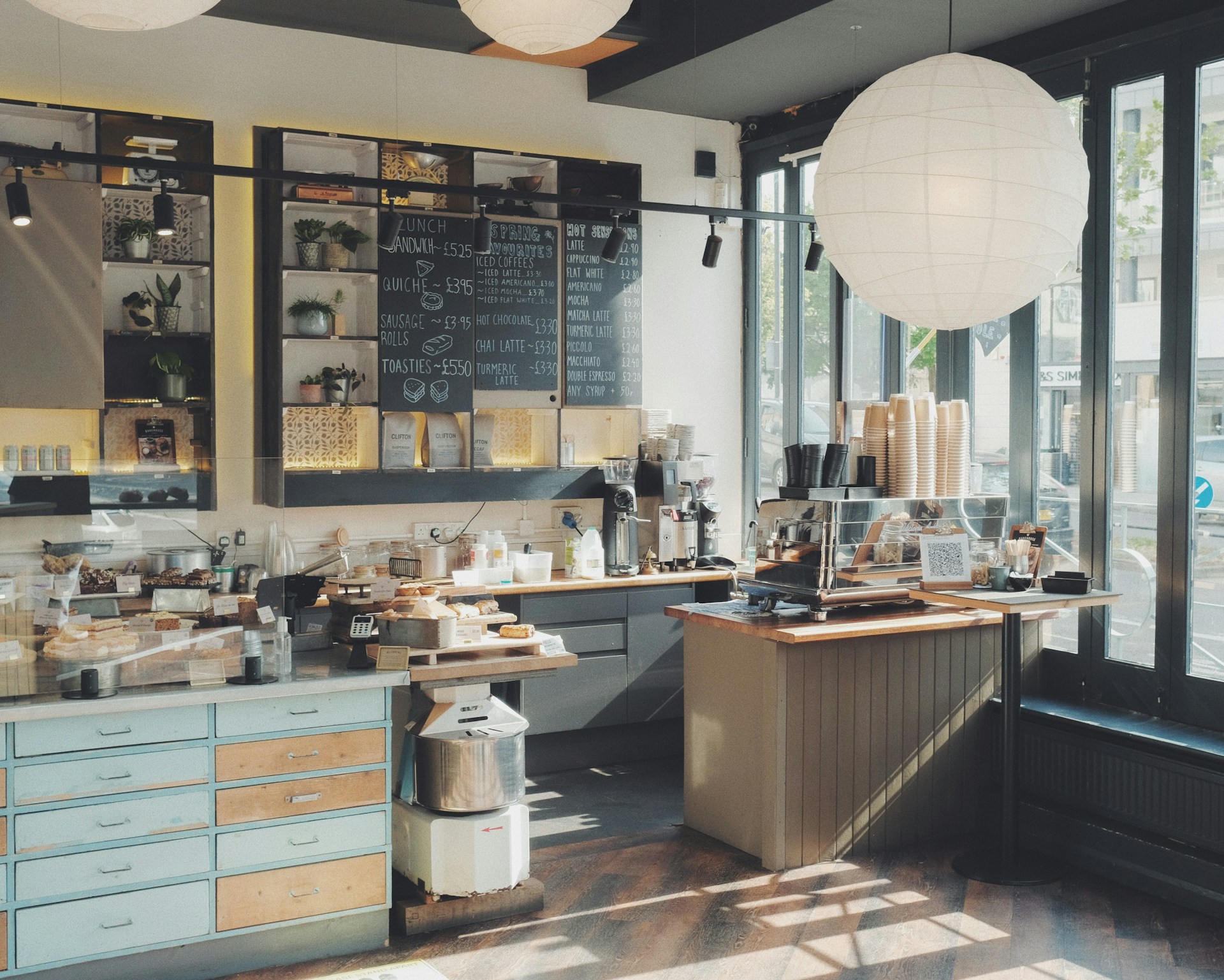 A cozy coffee shop interior with large glass windows allowing natural light to flood the space. Shelves on the wall display plants and coffee bags. The menu is written on a blackboard. The counter is equipped with a coffee machine, stacks of cups, and an array of pastries displayed on wooden trays. The aesthetic is warm and inviting, with light wood accents and industrial lighting.