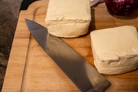 A wooden cutting board holds two large blocks of tofu, positioned near a sharp kitchen knife. In the background, part of a red onion is visible, adding contrast to the composition.