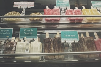 An assortment of gelato bars and other frozen treats are displayed behind a glass counter. Labels indicate flavors such as Belgian Chocolate, French Vanilla, and Tiramisu. Vegan options, including Strawberry Sorbet, are also present. Brightly colored popsicles, cookies, and waffles can be seen.