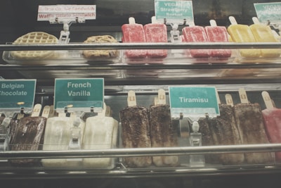 An assortment of gelato bars and other frozen treats are displayed behind a glass counter. Labels indicate flavors such as Belgian Chocolate, French Vanilla, and Tiramisu. Vegan options, including Strawberry Sorbet, are also present. Brightly colored popsicles, cookies, and waffles can be seen.