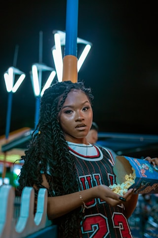 A person with braided hair is holding a box of popcorn while wearing a sports jersey. The setting appears to be outdoors at night, with bright streetlights and a casual, relaxed atmosphere.